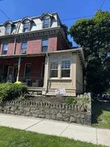 Historical red brick row house with a mansard roof, small sunroom addition, and a stone retaining wall next to a sidewalk.